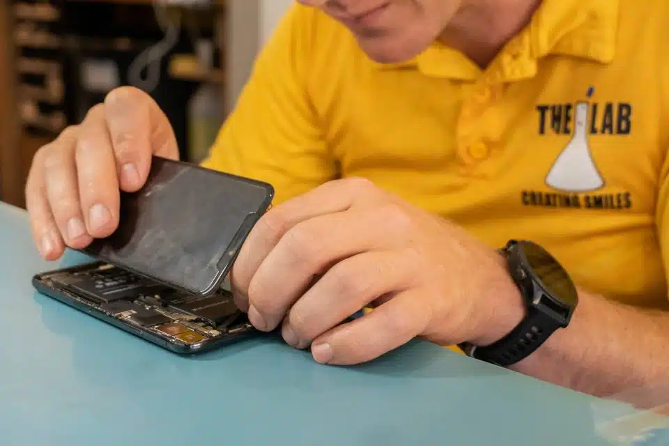 Hands opening a damaged smartphone on a blue countertop by Lyle wearing a yellow polo shirt with The Lab logo.