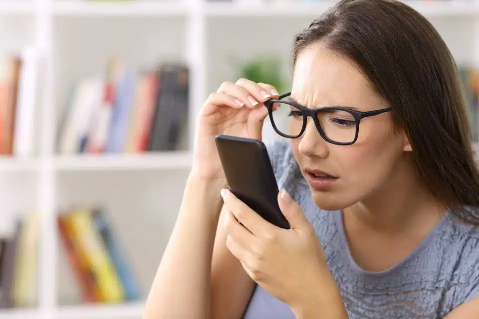 Woman squinting at her smartphone while holding the side of her glasses