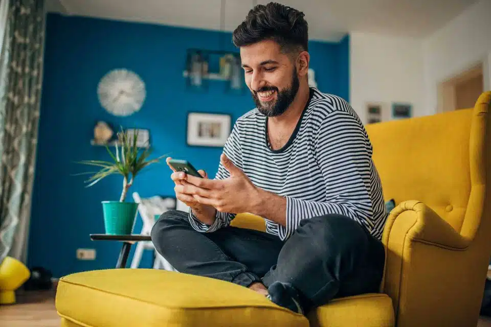 Bearded man in striped shirt sitting on a yellow chair smiling at the smartphone he holds in his hands