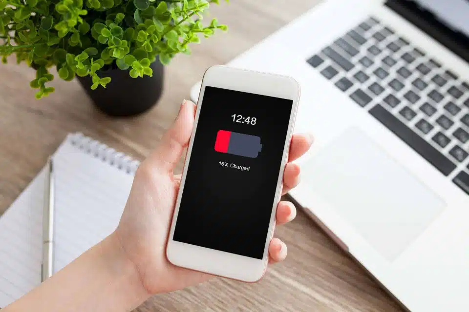 Hand holding a mobile phone showing a low battery next to a laptop and plant