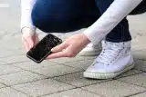 Person crouched near the ground after picking up their phone that has been dropped and has a cracked screen