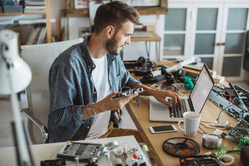 Man repairing the interior of an electronic device sitting at a desk while looking up information on a laptop