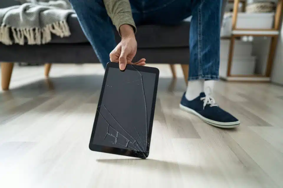 Hand holding a tablet leaning on the floor with a cracked screen next to their foot
