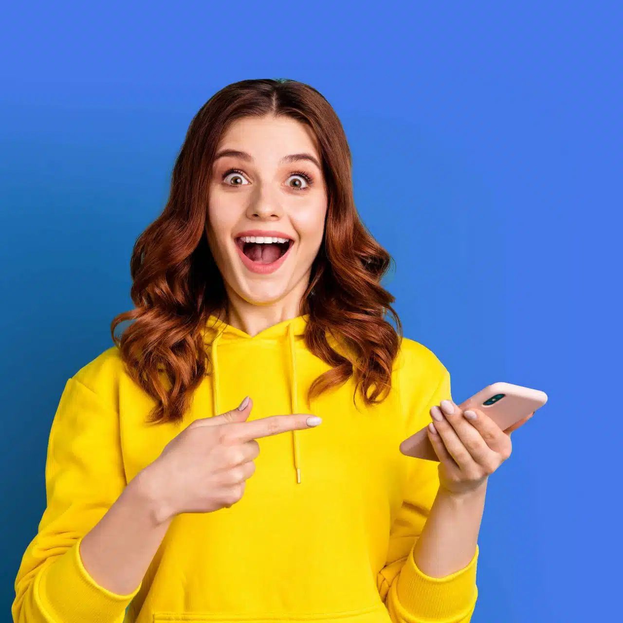 Woman with wavy brown hair and a yellow hoodie smiles as she points to the smartphone she's holding