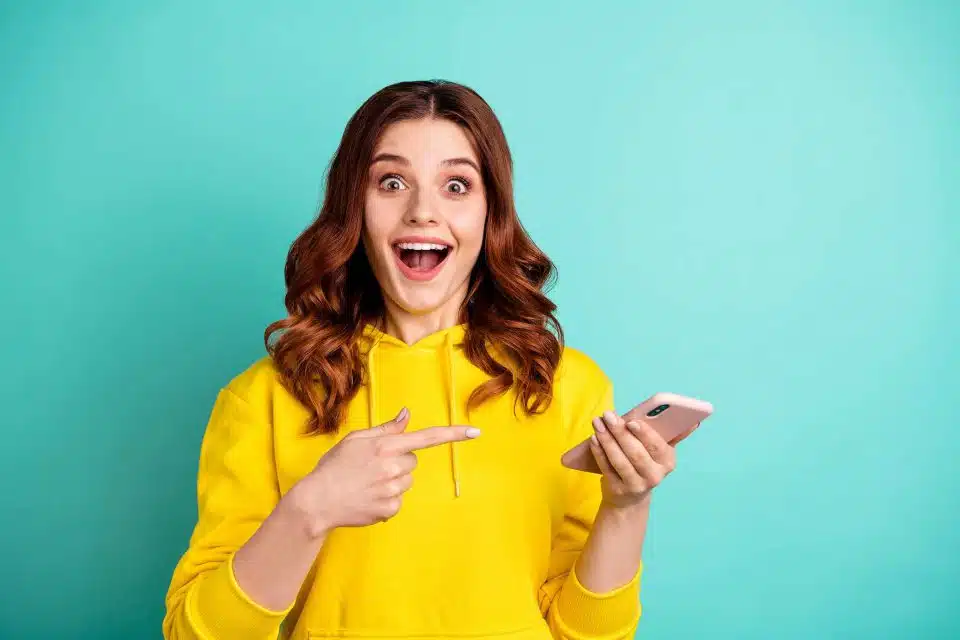 Brunette woman in a yellow hoodie pointing to a smartphone with an open-mouthed smile against a light teal background
