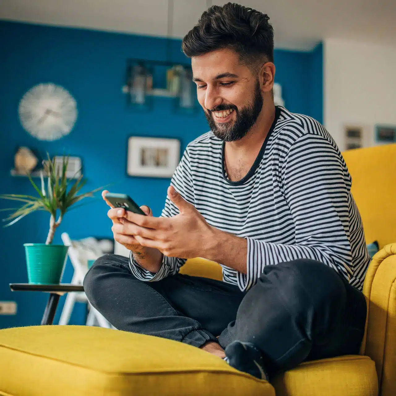 Bearded man in striped shirt sitting on a yellow chair smiling at the smartphone he holds in his hands
