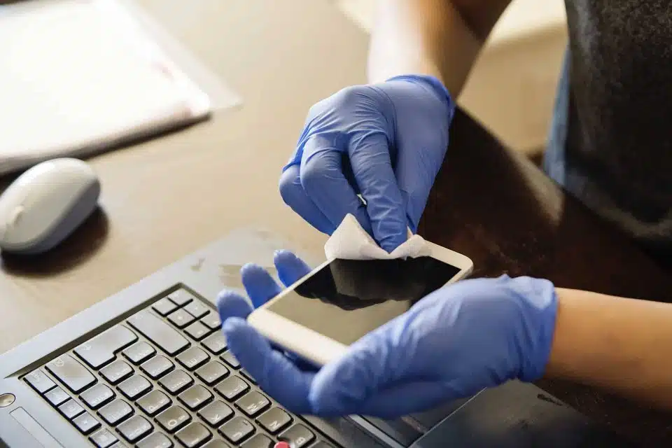 Hands in blue latex gloves using a cleaning wipe to clean the screen and surface of an iPhone