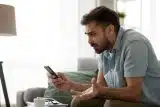 Brunette man with a beard holding his phone and looking at it in frustration